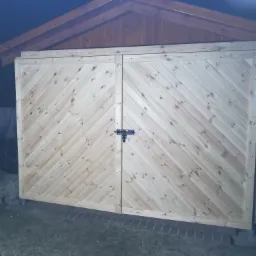 Nowe, dwuskrzydłowe drewniane wrota do garażu lub pomieszczenia gospodarczego, z ukośnym wzorem desek, zabezpieczone zasuwką, widoczne w słabym oświetleniu.