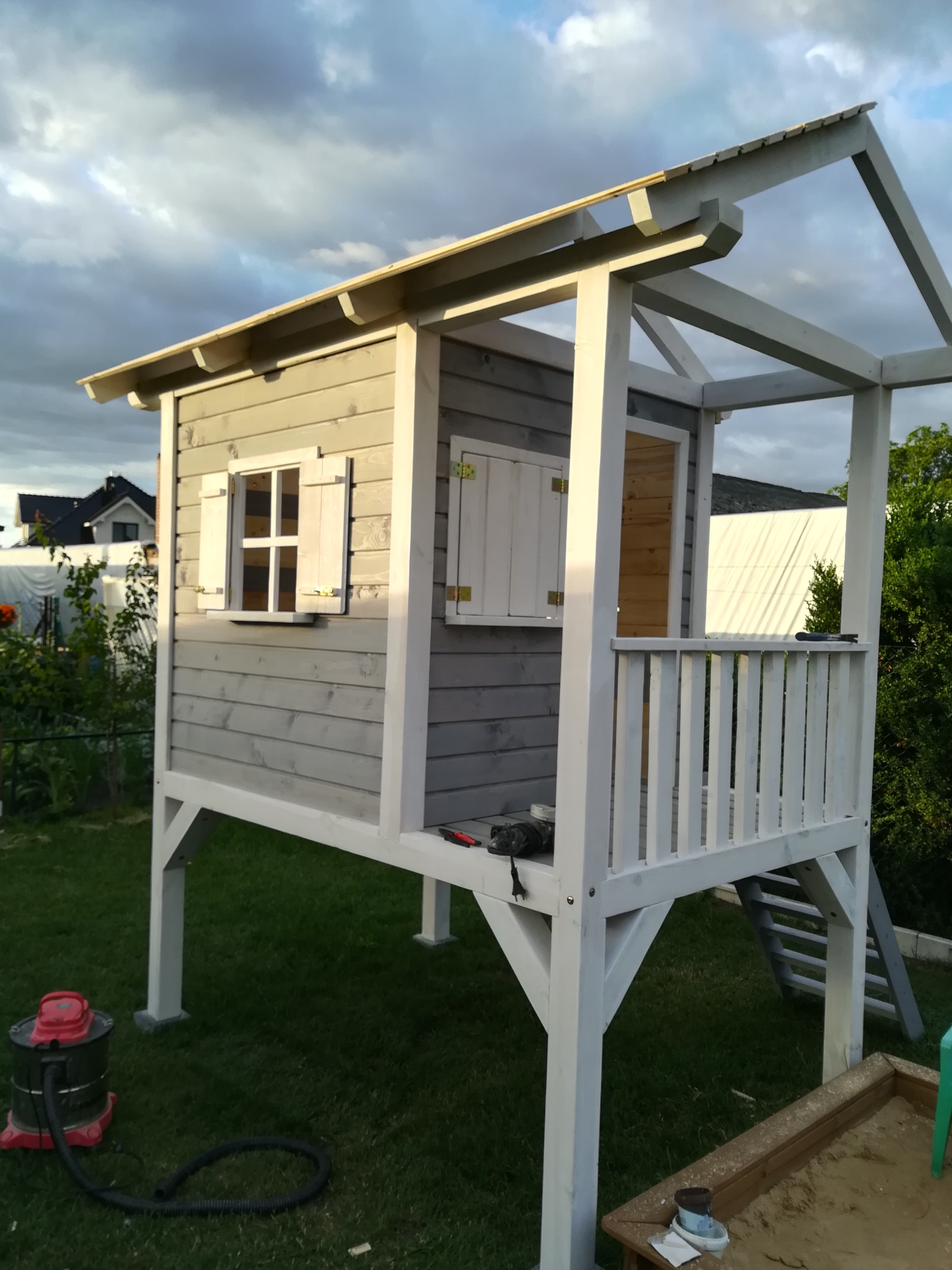 Szara, drewniana konstrukcja domku dla dzieci na podwyższeniu z białymi elementami wykończeniowymi, z otwartymi okiennicami, widoczny odkurzacz przemysłowy i piaskownica w ogrodzie.