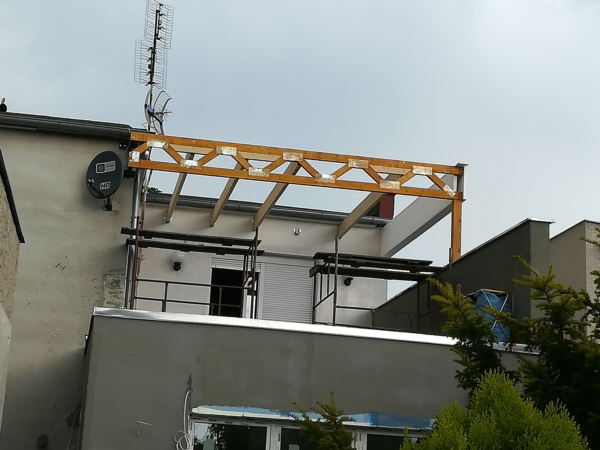 Drewniana konstrukcja z elementami metalowymi na dachu budynku z anteną satelitarną i telewizyjną, widok z dołu na szarą elewację w pochmurny dzień.