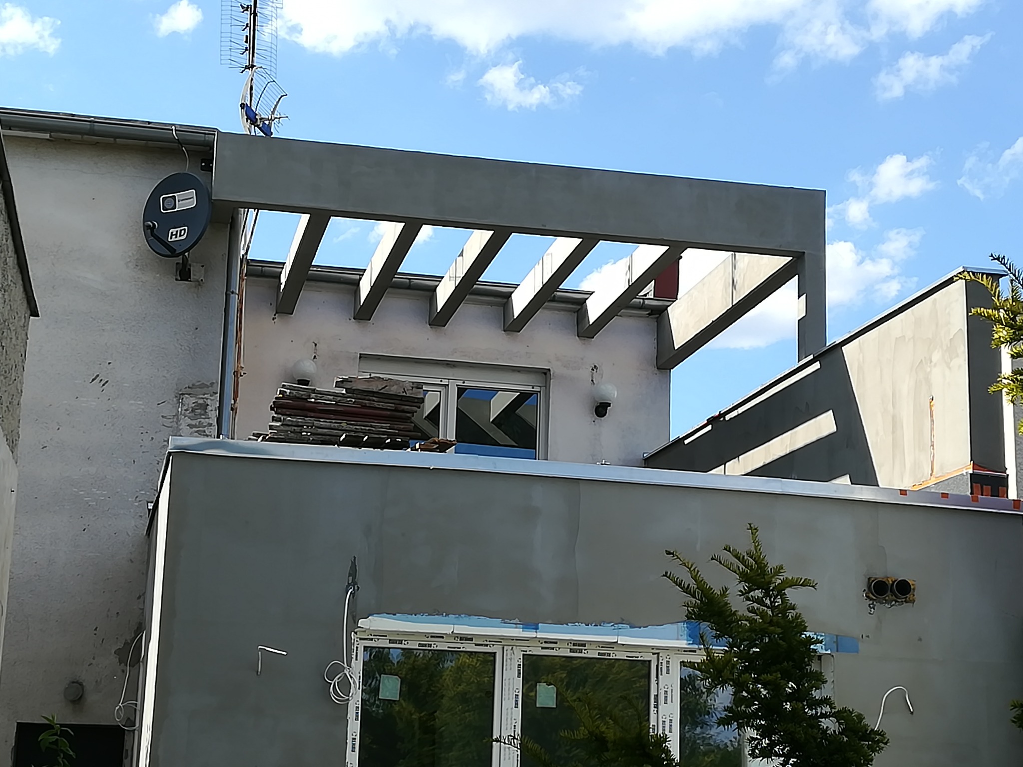 Szary budynek z nową elewacją i drewnianą konstrukcją zadaszenia nad tarasem, widoczna antena satelitarna na ścianie i stos dachówek przy oknie.