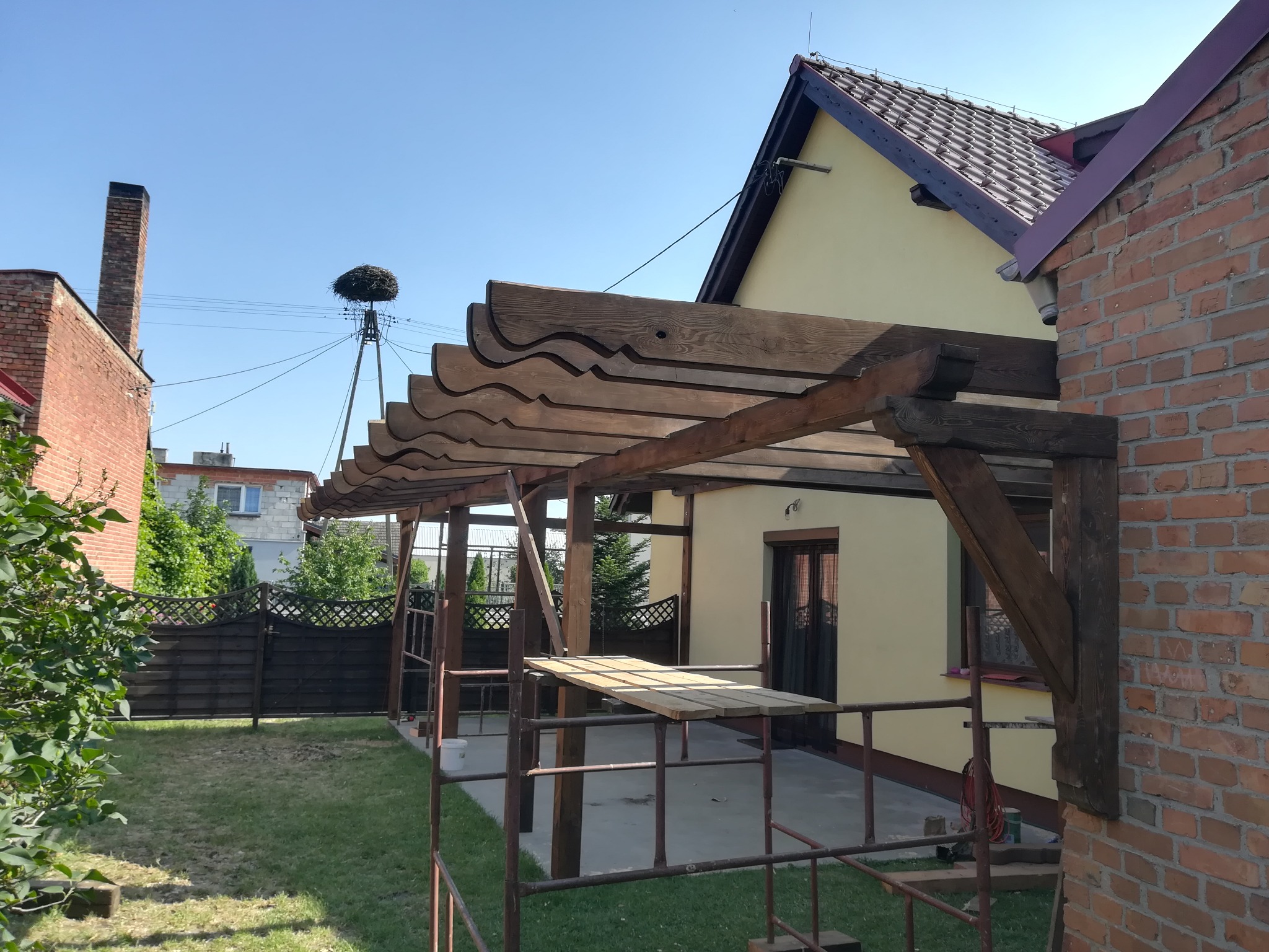 Drewniana pergola z falistym wykończeniem, w trakcie budowy, widoczna z rusztowania na tle domu z cegły i żółtą elewacją, na drugim planie gniazdo bocianie na słupie.