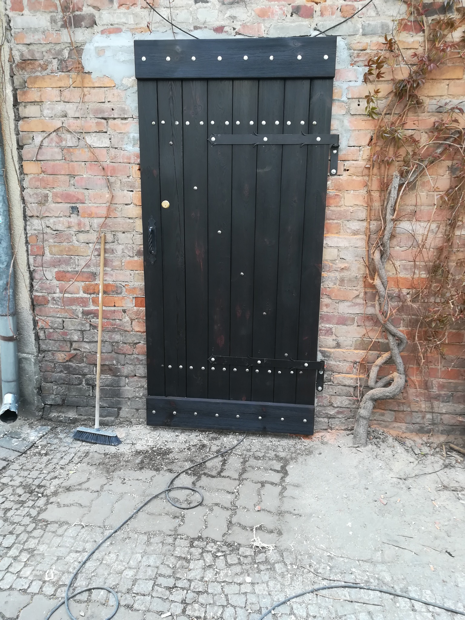 Nowo zamontowane, czarne, drewniane drzwi z ozdobnymi nitami w czerwonej ceglanej ścianie z pnącą roślinnością, obok stoi miotła.