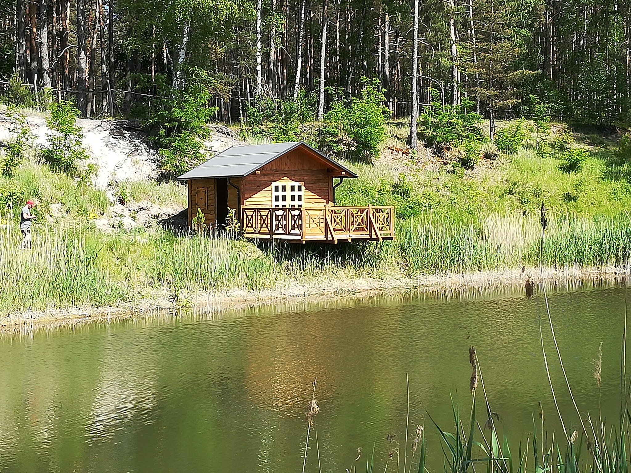 Drewniany domek letniskowy z małym tarasem nad brzegiem jeziora, otoczony lasem, z widocznym odbiciem w wodzie i osobą w oddali.