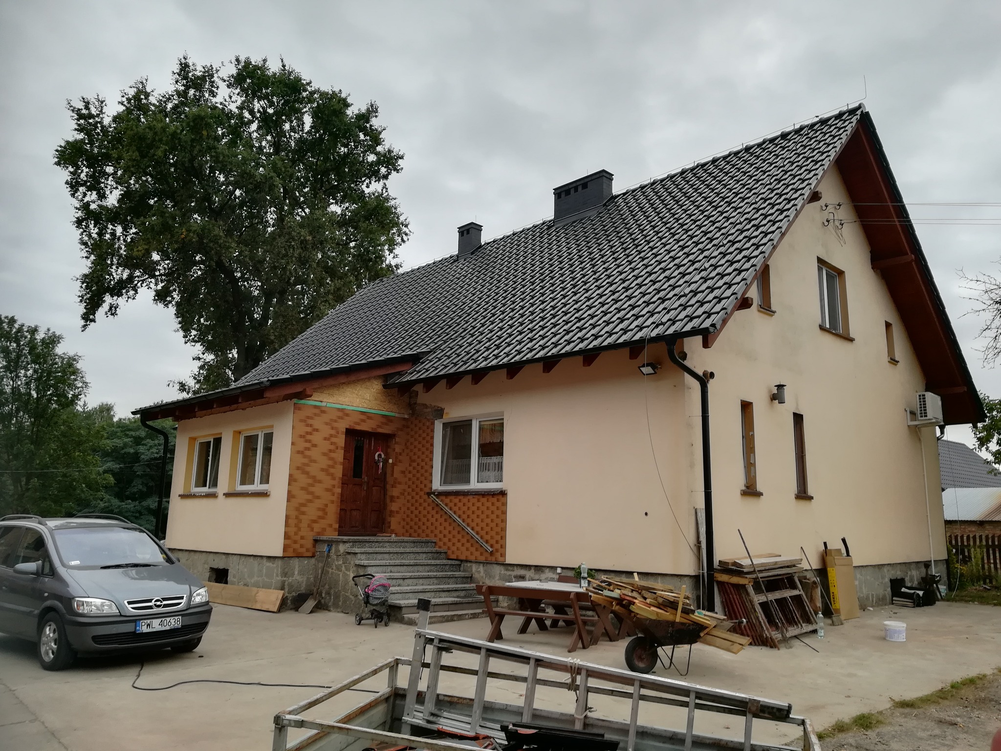 Dom jednorodzinny z nowym, ciemnoszarym dachem z dachówki ceramicznej, widoczne obróbki blacharskie, rynny, fragment ocieplonego poddasza i elementy elewacji po remoncie, na podwórku częściowo...