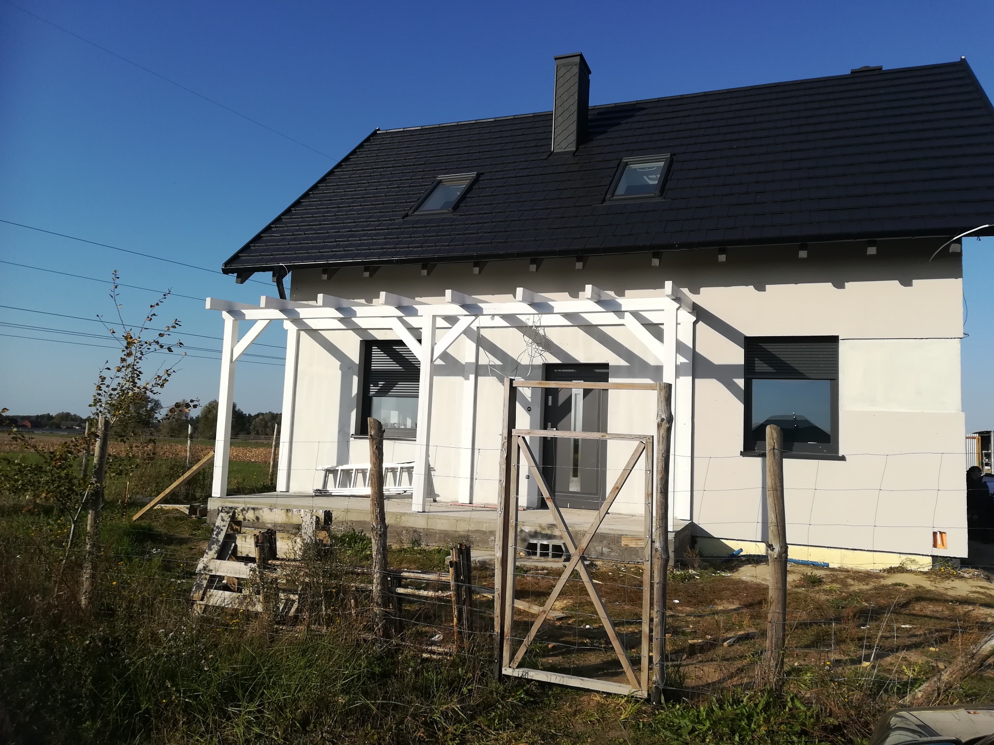 Dom jednorodzinny z czarnym dachem z dachówki, białymi oknami i białą pergolą, widoczny zza prowizorycznego ogrodzenia z siatki.