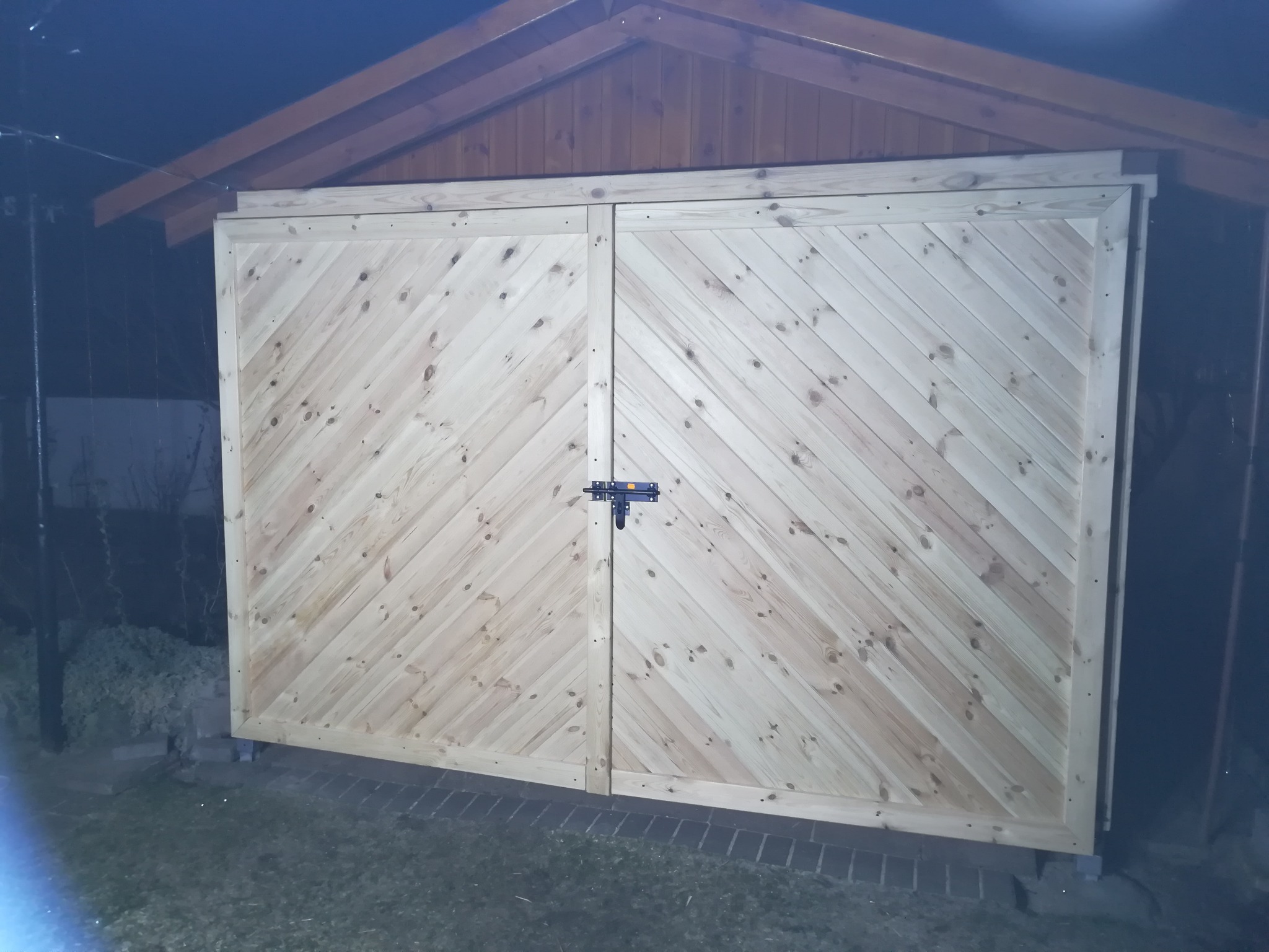 Nowe, dwuskrzydłowe drewniane wrota do garażu lub pomieszczenia gospodarczego, z ukośnym wzorem desek, zabezpieczone zasuwką, widoczne w słabym oświetleniu.