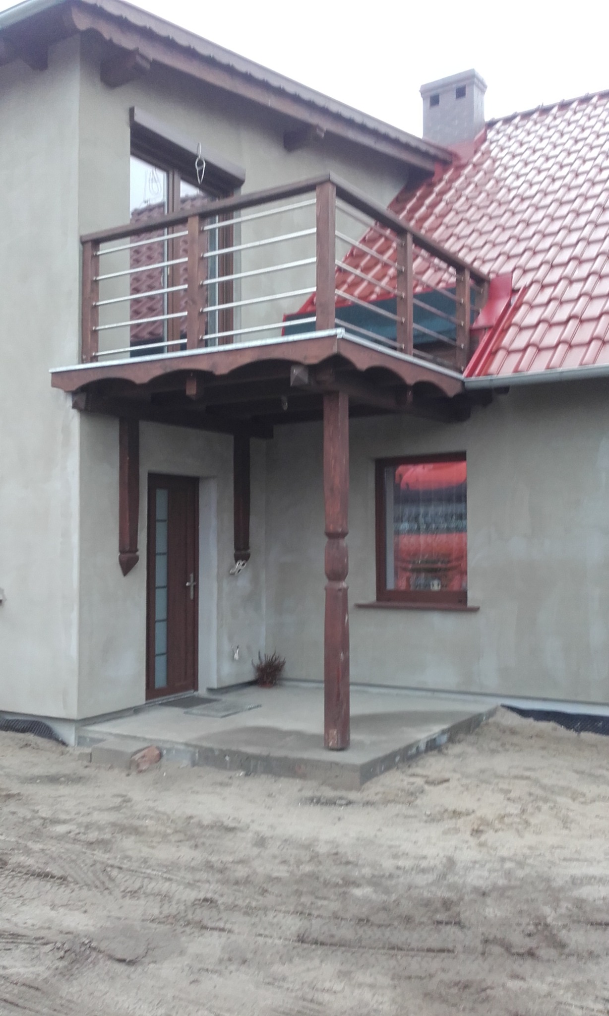 Wykończony dom z czerwonym dachem, drewnianym balkonem z metalową balustradą i podcieniem wspartym na drewnianych kolumnach. Uwagę zwraca świeżo położona, błyszcząca dachówka. Teren wokół domu...