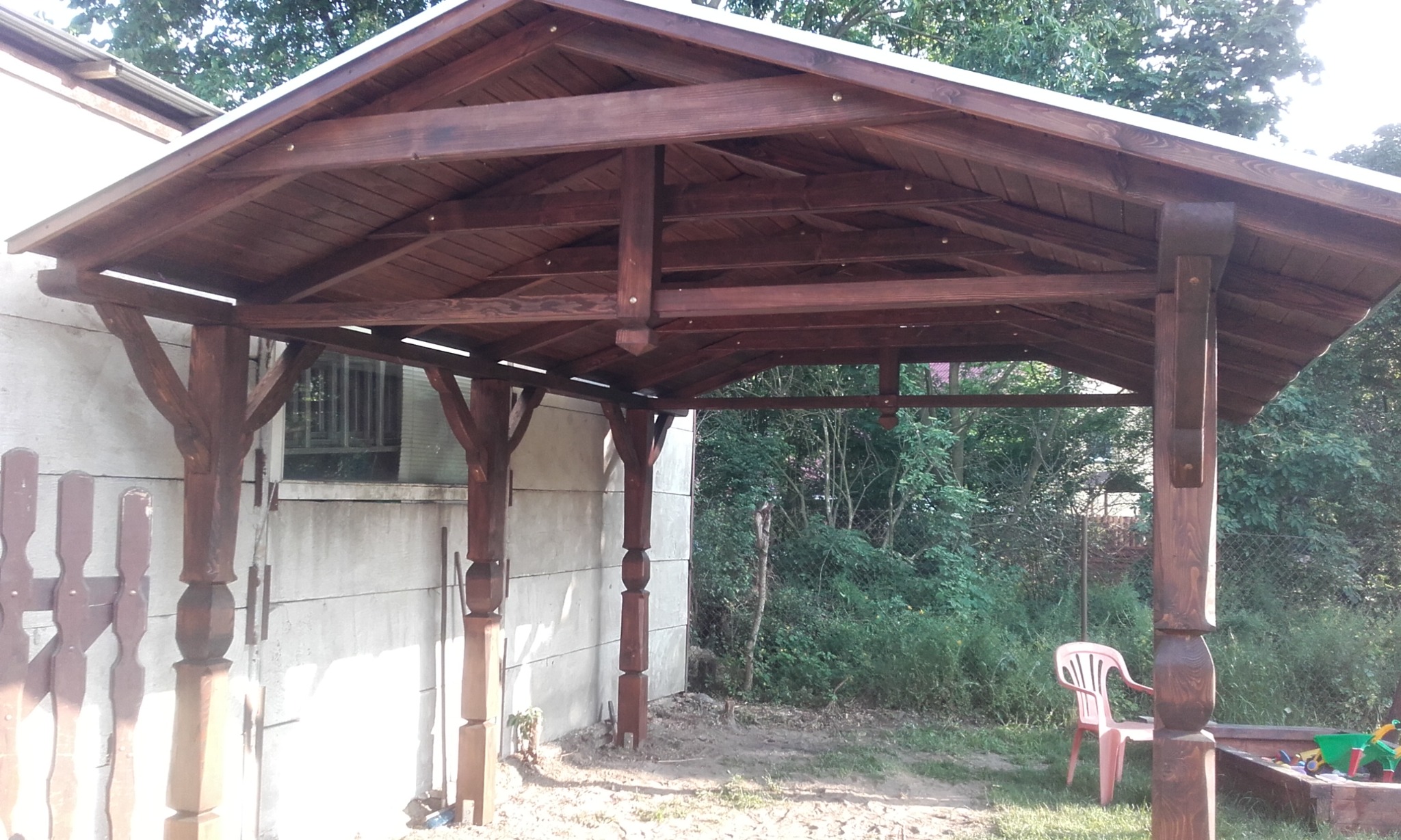 Drewniana altana ogrodowa z ciemnobrązowymi belkami i białym dachem, widok z zewnątrz na tle zieleni, obok piaskownica i plastikowe krzesło.