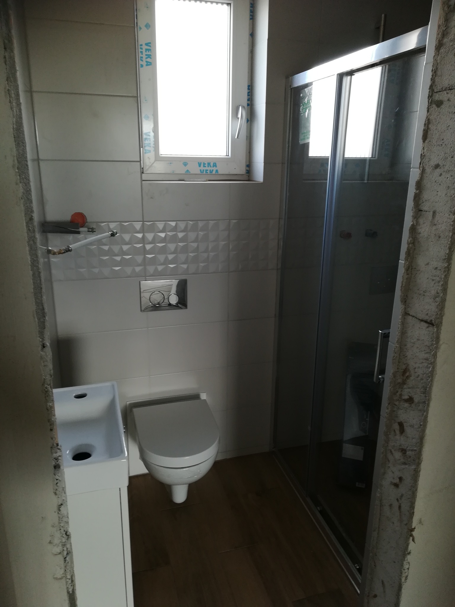 Wnętrze łazienki w trakcie remontu: białe płytki z wzorem, podwieszana toaleta, umywalka z szafką, kabina prysznicowa z przesuwnymi drzwiami i okno z ramą PCV.