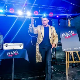 Mężczyzna w złotym fraku prezentuje trik z połączonymi metalowymi obręczami na scenie z oświetleniem i logiem 'MAGO Magic Show' w tle.