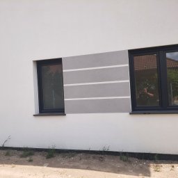 Lis Docieplenia Dachy - Fragment elewacji budynku z dwoma oknami w czarnych ramach, szara dekoracja z białymi pasami, widoczne odbicie fotografa w szybie.