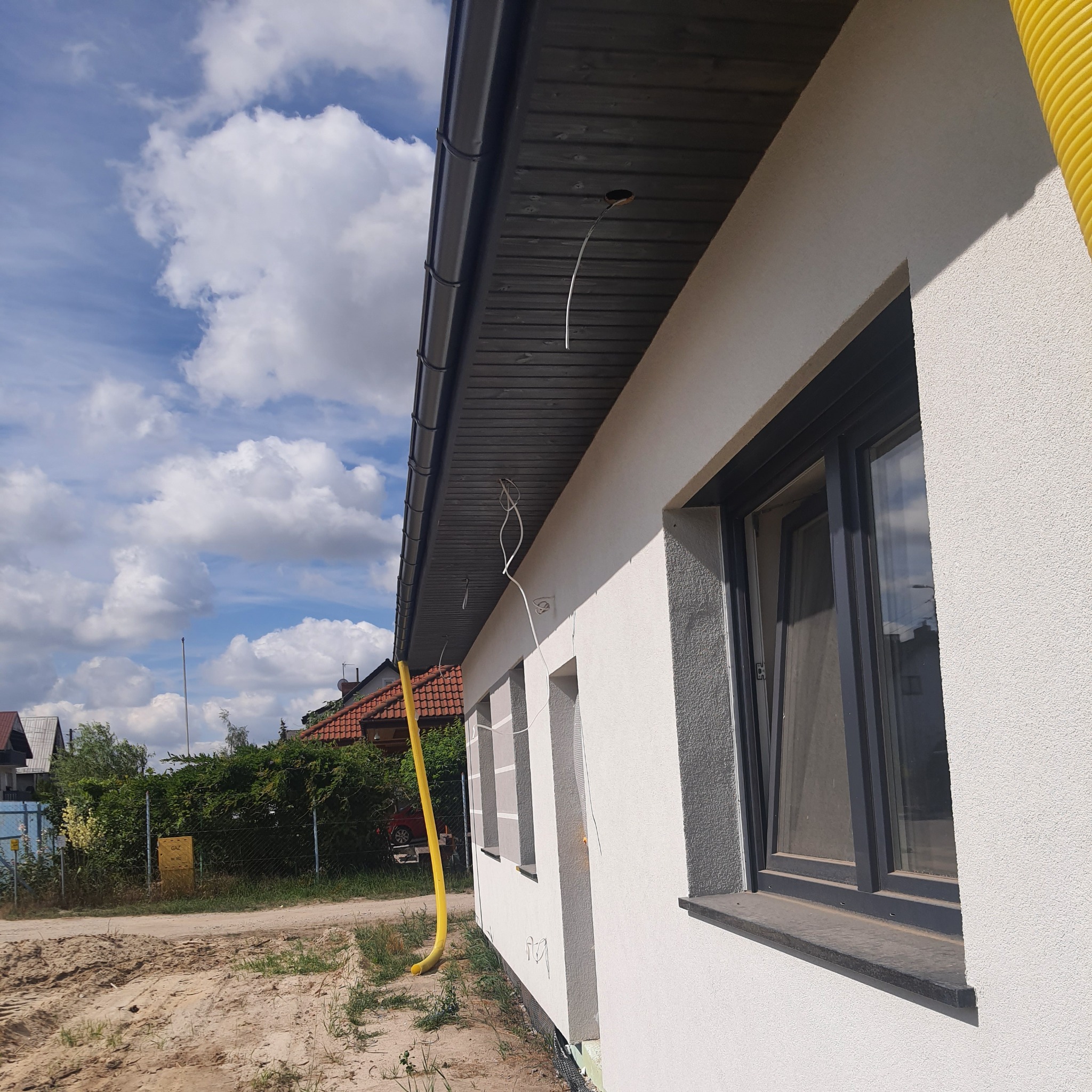 Fragment ściany budynku z nową, jasną elewacją o strukturze baranka, ciemne okna, czarna podbitka dachowa, wystające przewody i żółta rura wentylacyjna przy elewacji na tle błękitnego nieba...