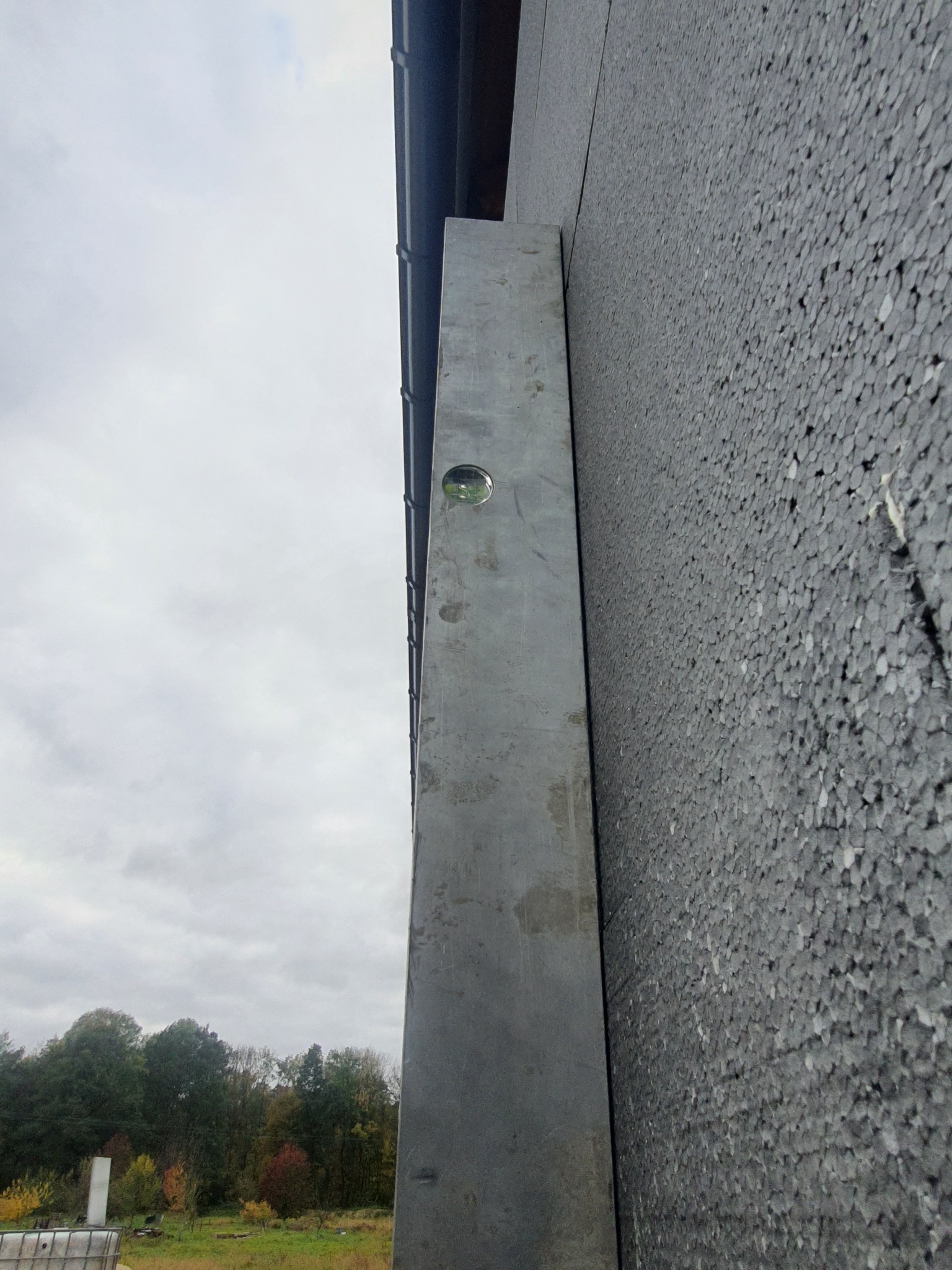 Szara ściana budynku pokryta styropianem z zamontowanym metalowym profilem pionowym, widoczna poziomica, tło stanowi pochmurne niebo i drzewa.