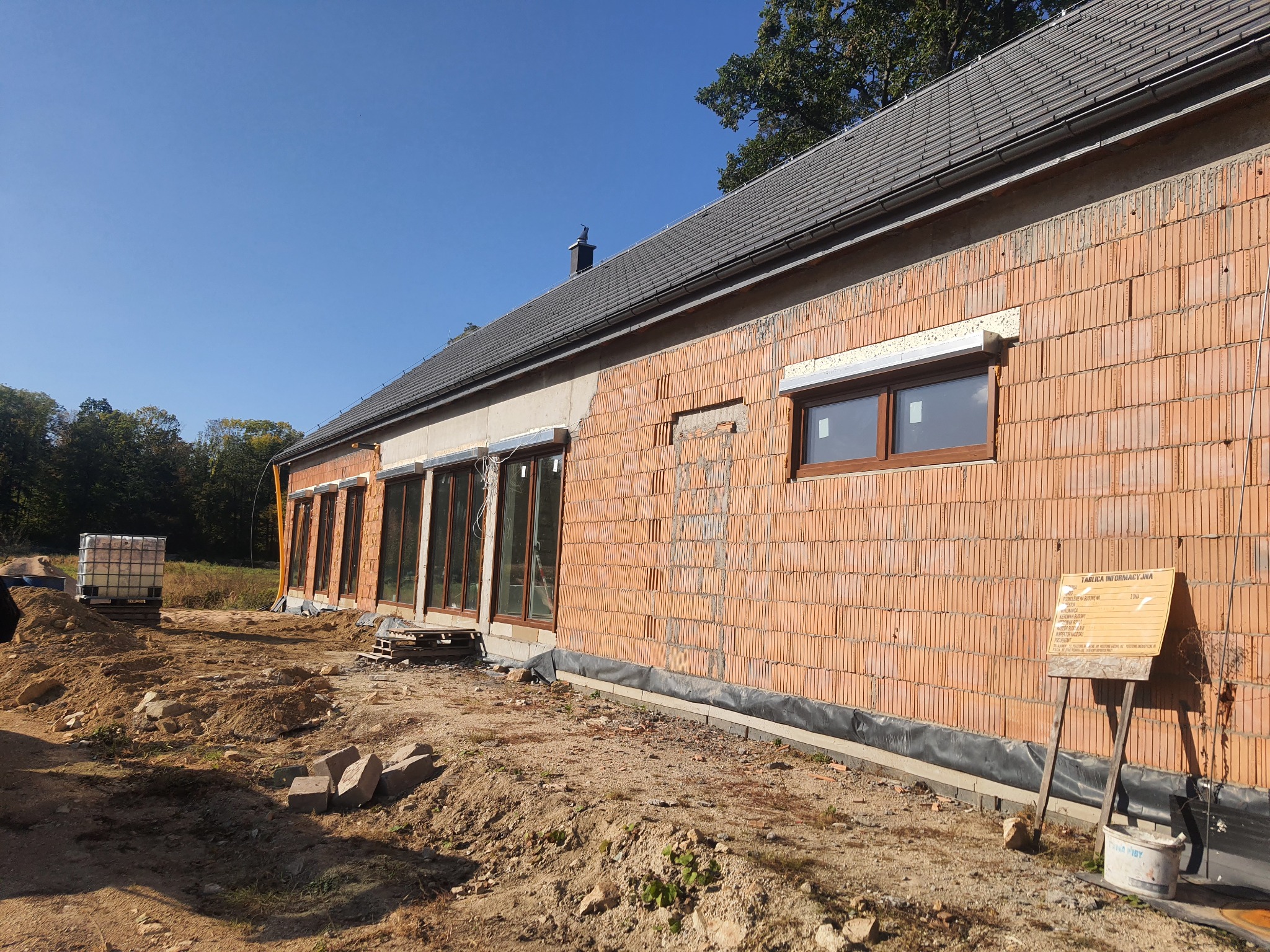 Budowa domu jednorodzinnego w trakcie realizacji: widoczne ściany z czerwonej cegły, okna z brązowymi ramami, częściowo otynkowana elewacja, dach z szarej dachówki, teren budowy z hałdami ziemi...