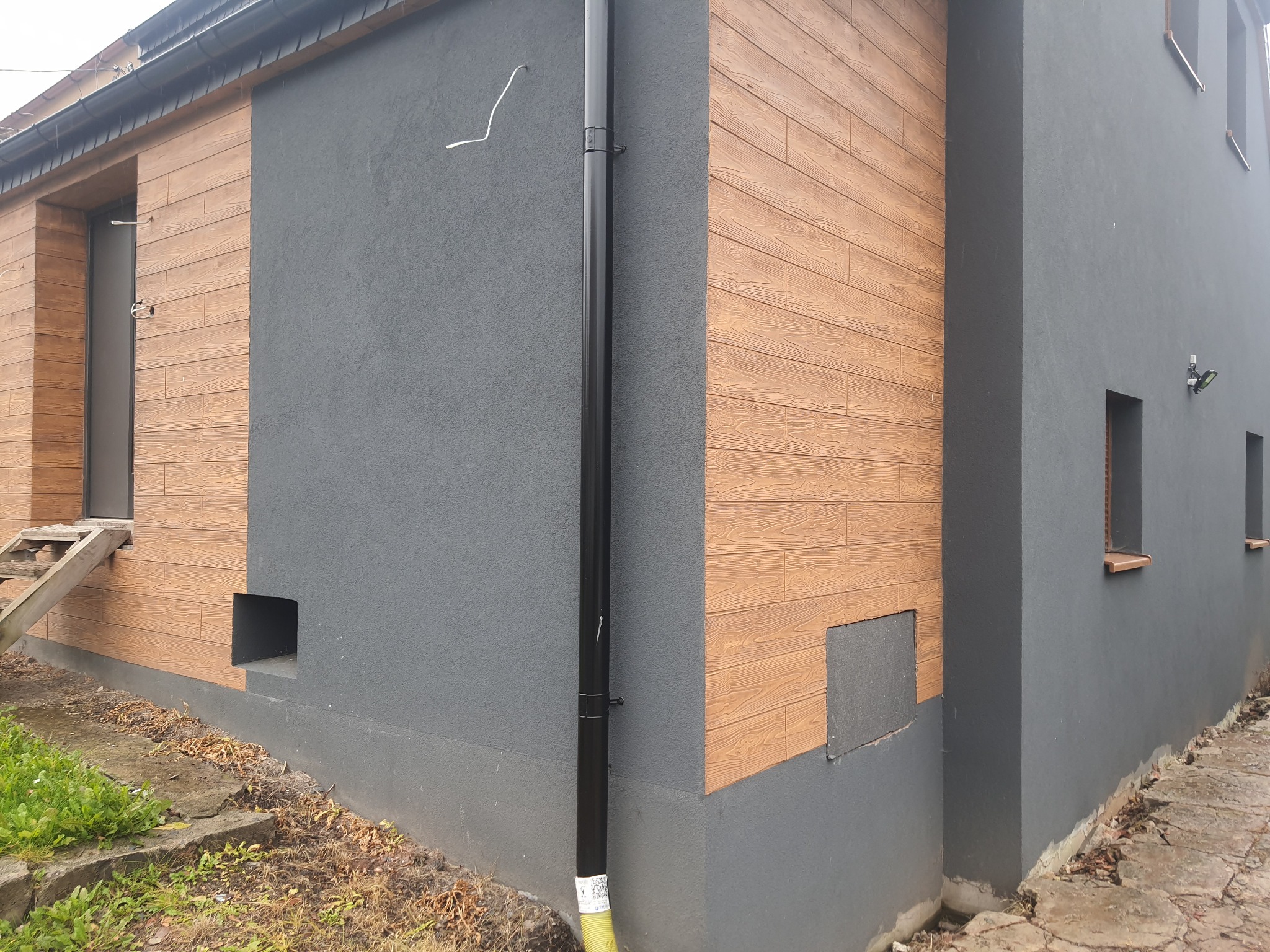 Szary budynek z drewnopodobną elewacją, widoczne rynna, okna i drewniane schody wejściowe.