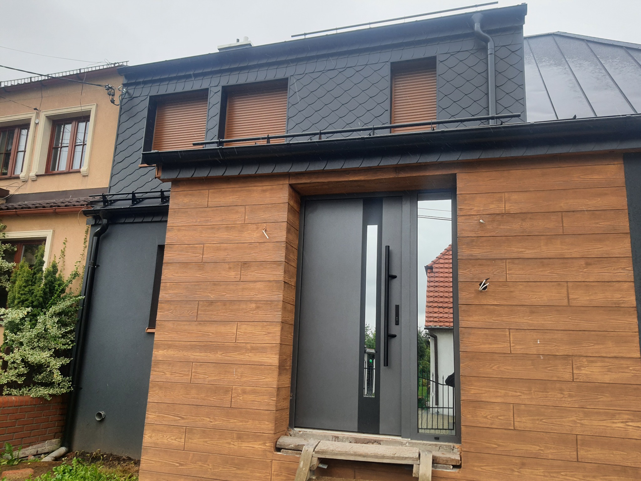 Nowoczesny dom z elewacją imitującą drewno, grafitową stolarką okienną i drzwiową oraz dachem pokrytym łupkiem, widoczne niedokończone prace wykończeniowe.