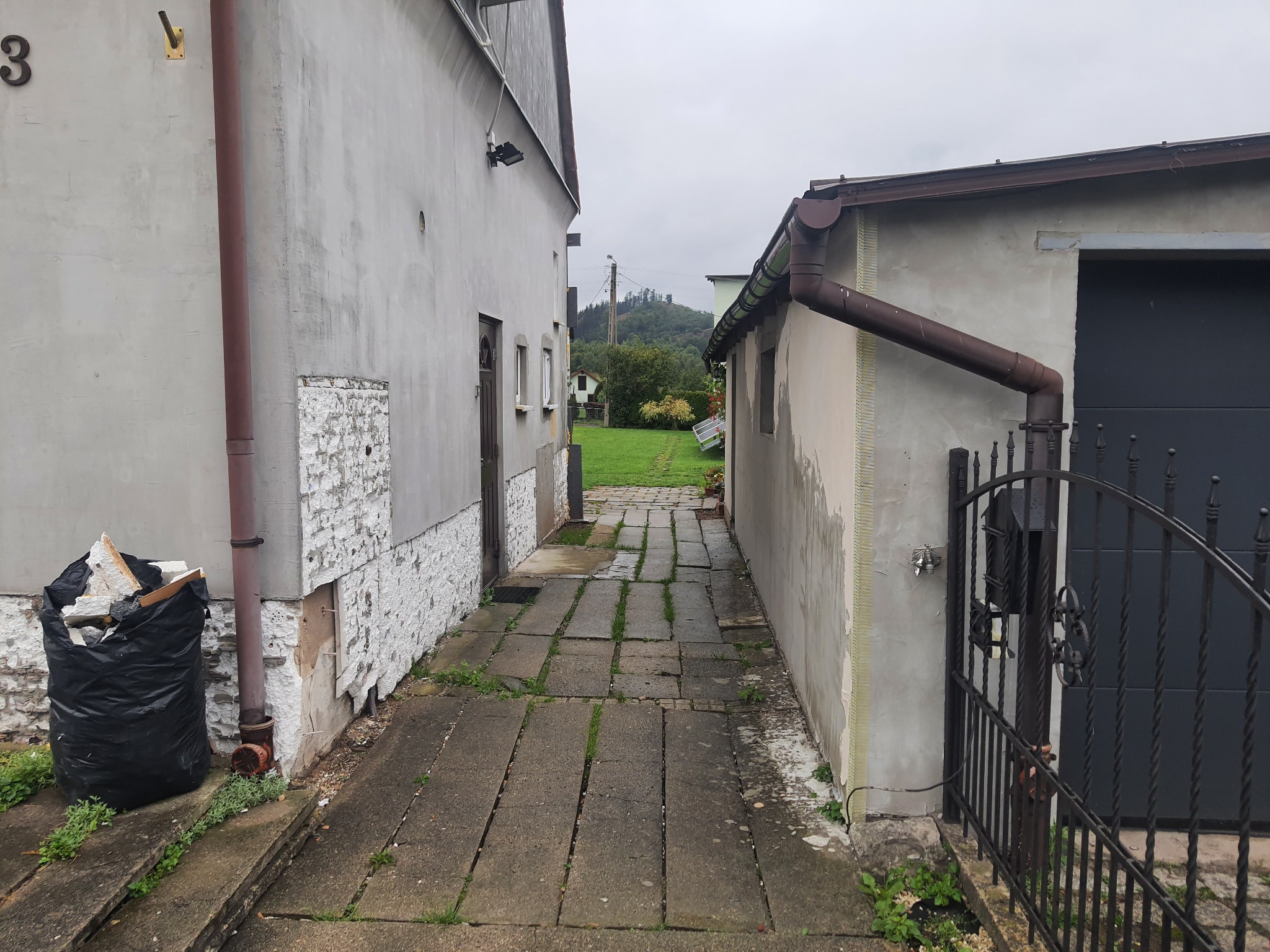 Wąskie przejście między dwoma budynkami z elewacją w trakcie remontu, widoczny czarny worek ze śmieciami budowlanymi i odsłonięta izolacja, w tle zielony krajobraz.