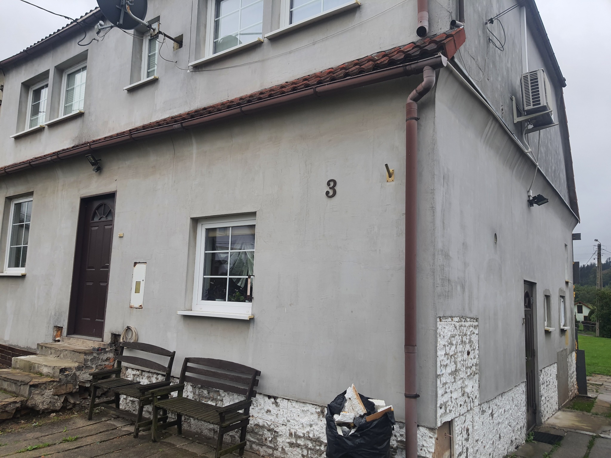 Szara elewacja domu z numerem 3 w Bydgoszczy, widoczne okna z białymi ramami, brązowe rynny i drzwi wejściowe, dwa drewniane krzesła przed wejściem, ślady wcześniejszych prac na elewacji.
