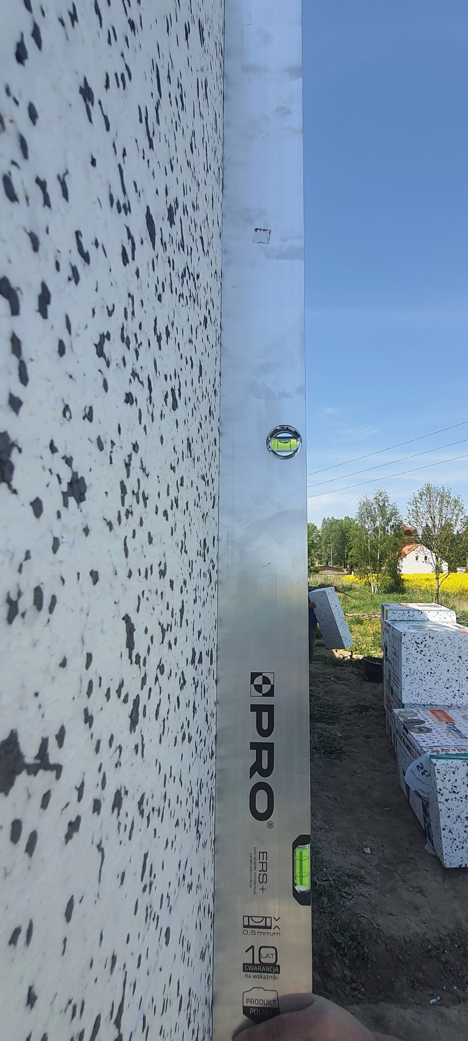 Sprawdzanie poziomu ściany ocieplonej styropianem z fakturą 'baranka' za pomocą poziomicy PRO ERS+ z 10-letnią gwarancją na wskaźniki, w tle teren budowy i błękitne niebo.