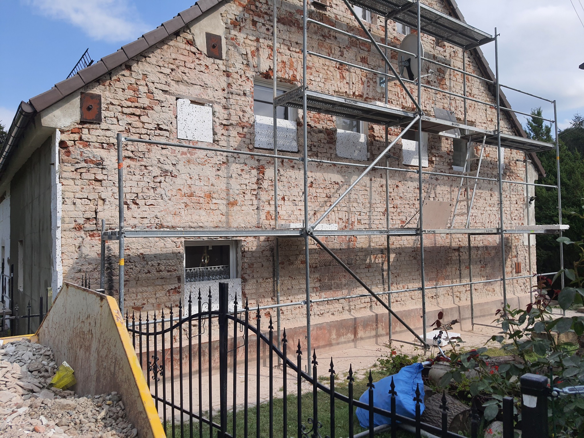 Dom z cegły w trakcie prac elewacyjnych, widoczne rusztowania, częściowo ocieplone okna styropianem, gruz w kontenerze na pierwszym planie.