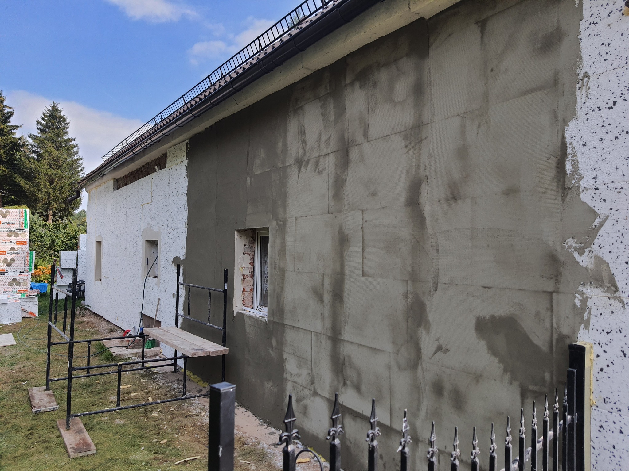 Ocieplanie ściany budynku: fragment z nałożoną warstwą kleju z siatką, fragment z izolacją styropianową, widoczne rusztowanie, materiały budowlane i ogrodzenie.