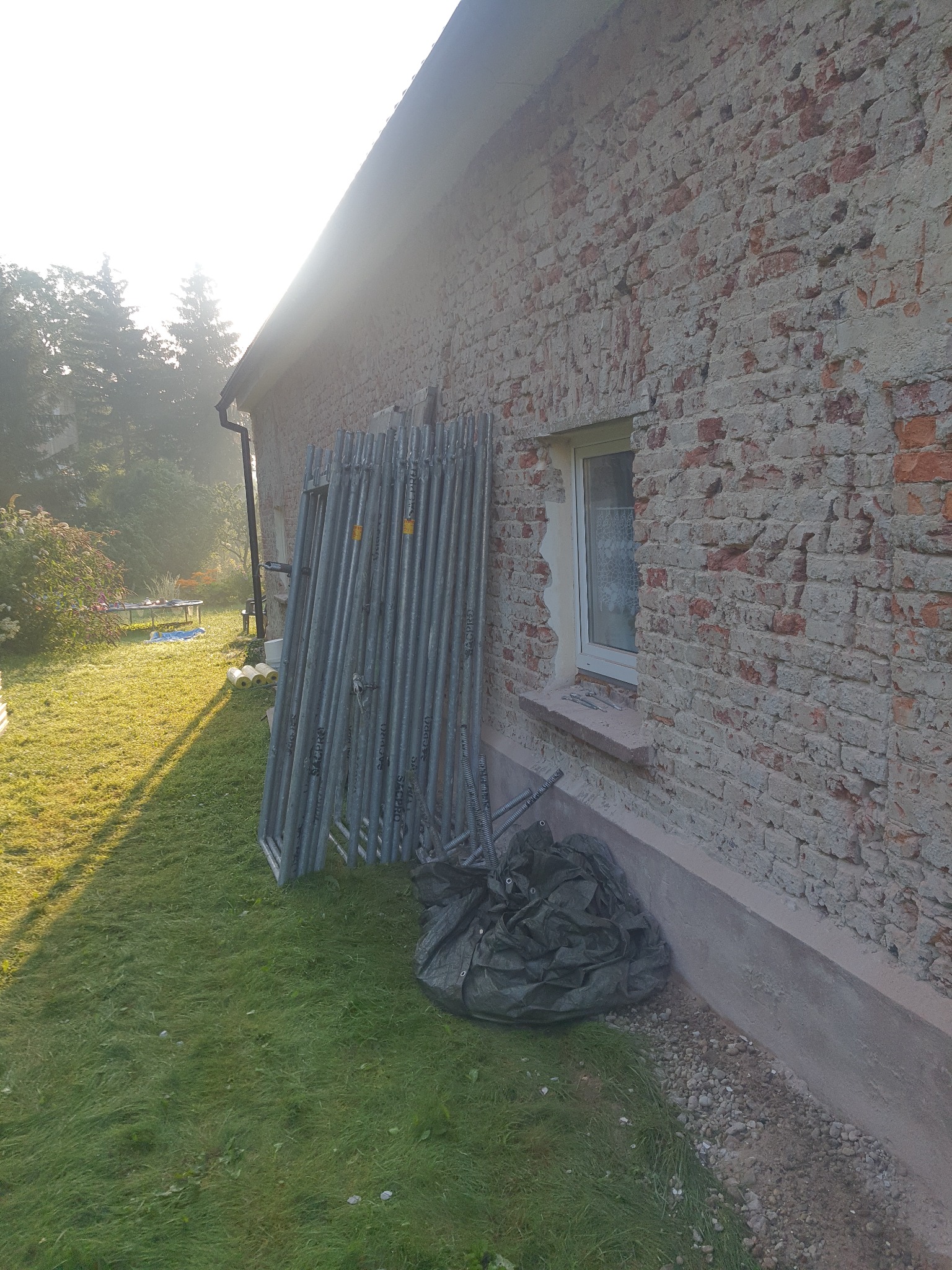 Stojące rusztowanie przy ceglanej ścianie domu z widocznymi śladami napraw, okno z firanką i workiem gruzu na tle trawnika i drzew w porannym słońcu.