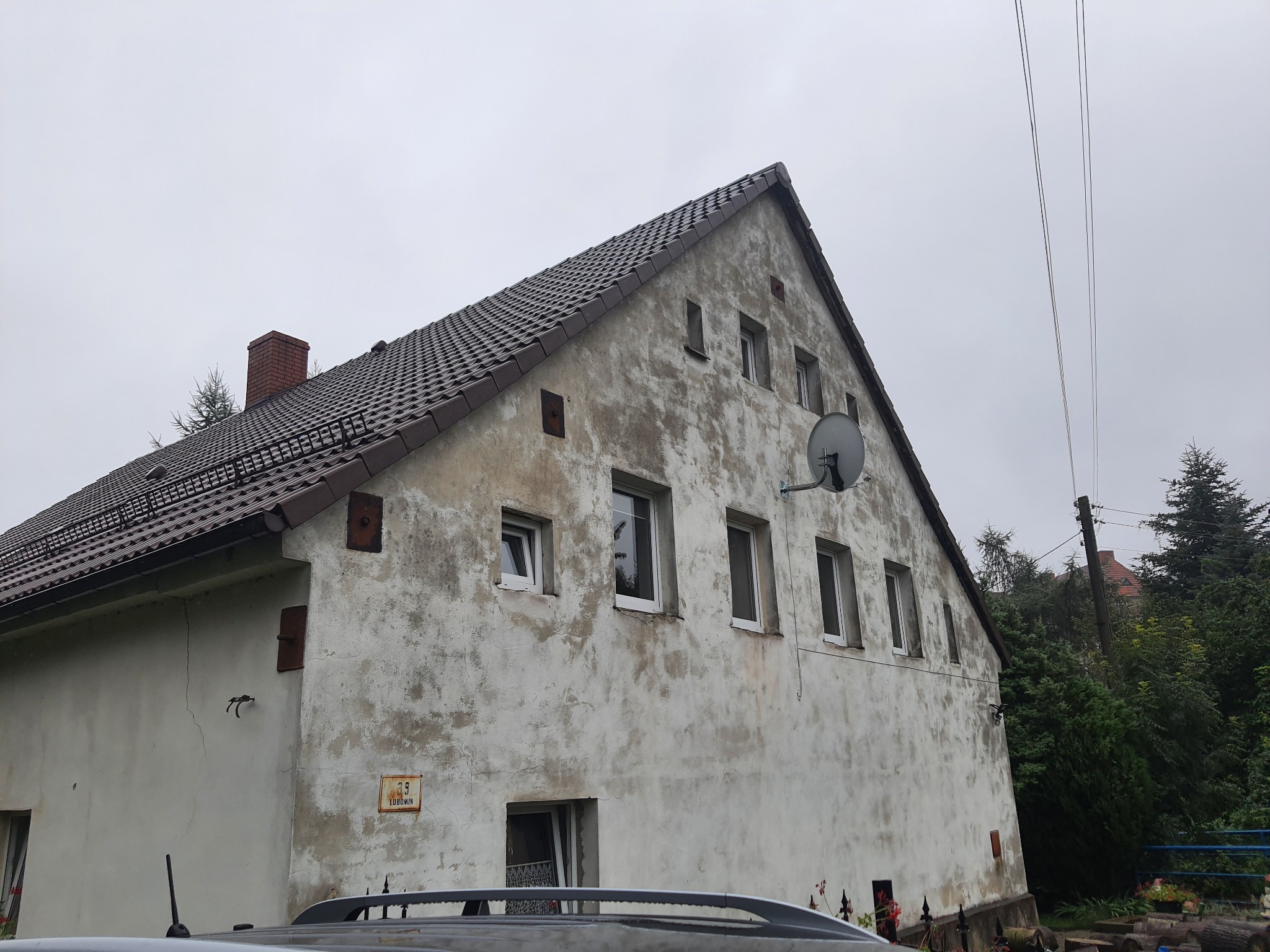 Stary, dwukondygnacyjny dom z brązową dachówką i popękaną, białą elewacją; widoczne okna, satelita, komin i tabliczka z numerem domu.