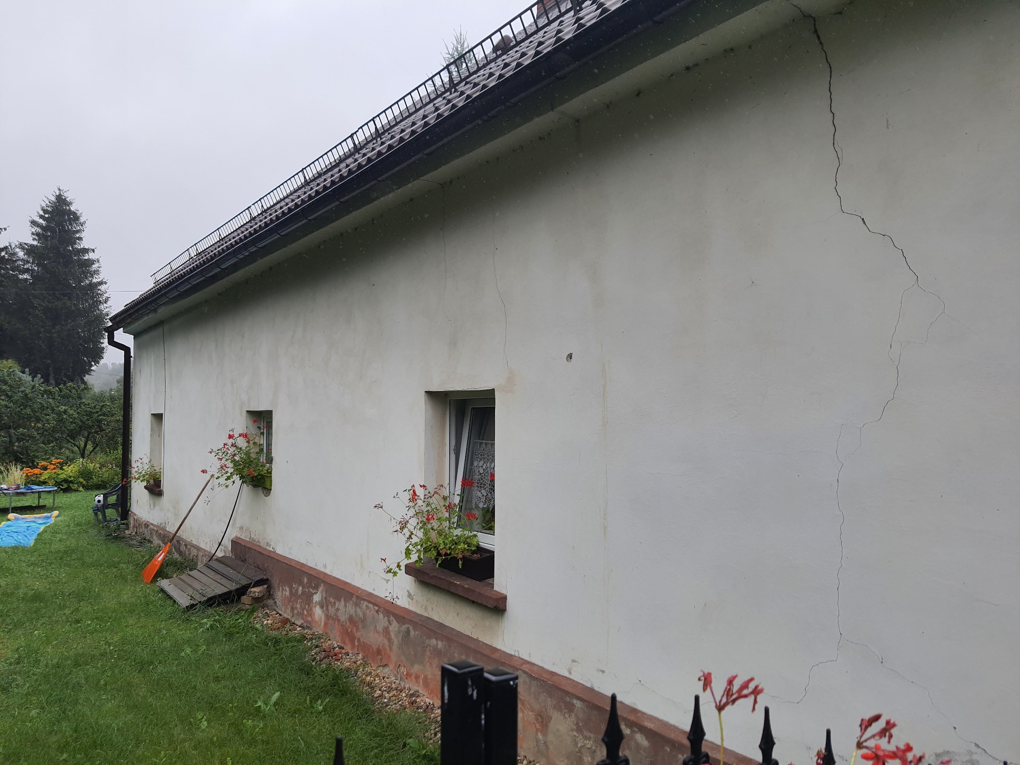 Boczna ściana domu z widocznymi pęknięciami na elewacji, dwa okna z czerwonymi kwiatami w doniczkach, drewniany podest i pomarańczowa łopata oparta o ścianę.