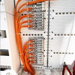 Dwa panele krosownicze z podłączonymi pomarańczowymi kablami, uporządkowanymi za pomocą opasek zaciskowych, widoczne narzędzia: kombinerki z czerwonymi i żółtymi rękojeściami oraz szczypce tnące...