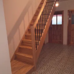 Jasne drewniane schody z czarnymi metalowymi balustradami, prowadzące do góry, z widoczną drewnianą ramą drzwi i wzorzystą podłogą w tle.