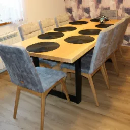 Jasny, drewniany stół z czarnymi, metalowymi nogami i ośmioma szarymi krzesłami tapicerowanymi, nakryty czarnymi podkładkami, z zabawką na środku, na tle wzorzystej tapety i drewnianej podłogi.