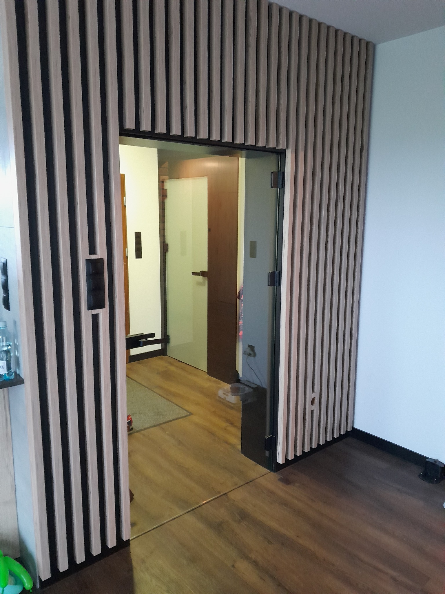 Szklane drzwi przesuwne z czarną ramą, otoczone drewnianą ścianką działową z pionowych listew, widok na wnętrze mieszkania z drewnianą podłogą.