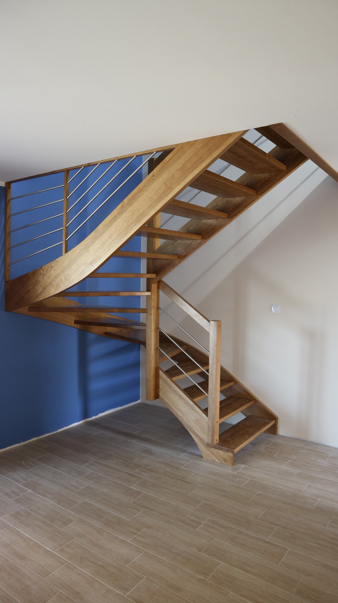 Drewniane schody spiralne z jasnego drewna z metalowymi poręczami, umieszczone w rogu pokoju z niebieską i białą ścianą, na podłodze płytki imitujące drewno.