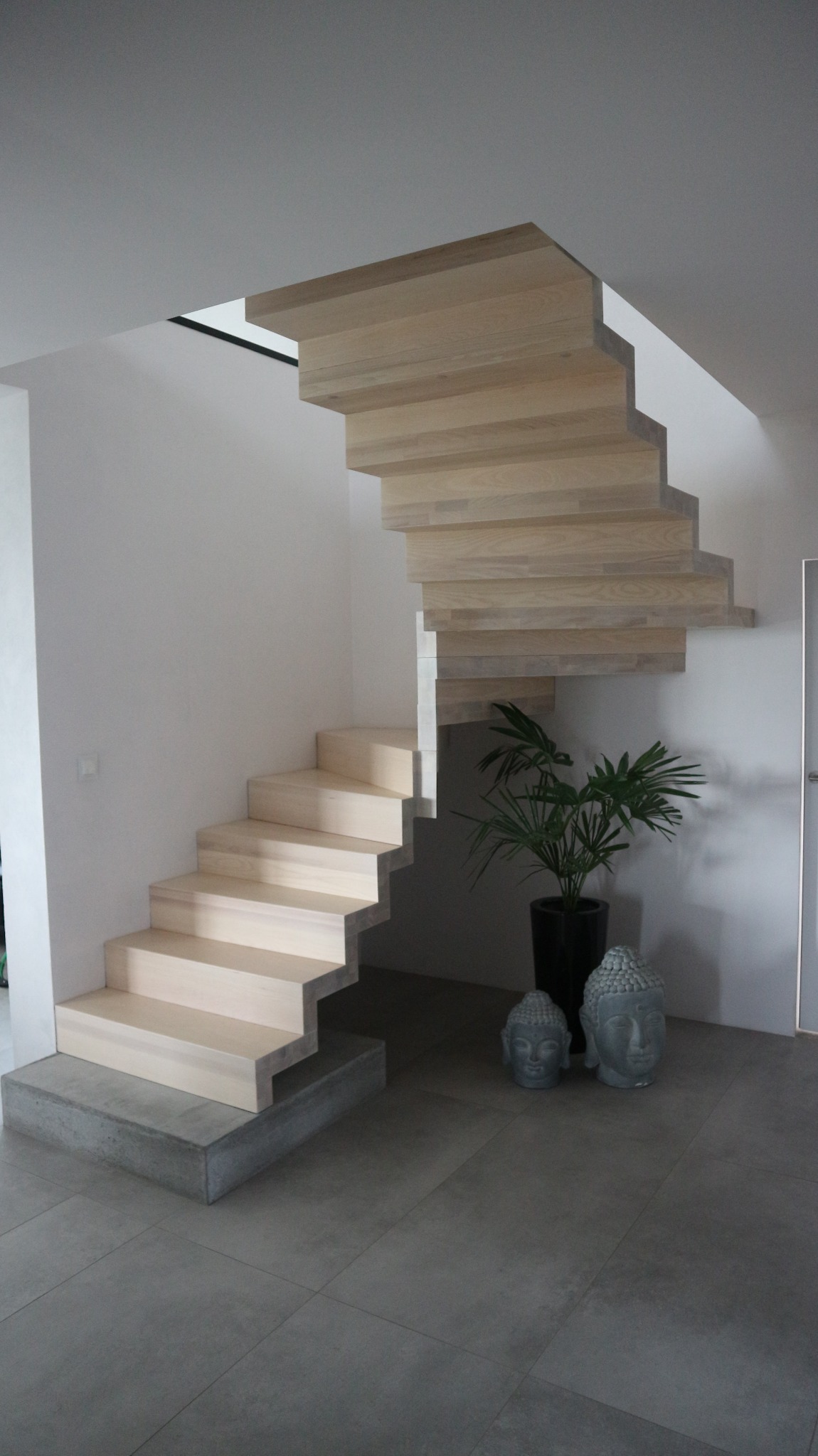 Nowoczesne, jasne, drewniane schody wiszące z betonową podstawą, uzupełnione rośliną doniczkową i dekoracyjnymi popiersiami Buddy na szarej podłodze.