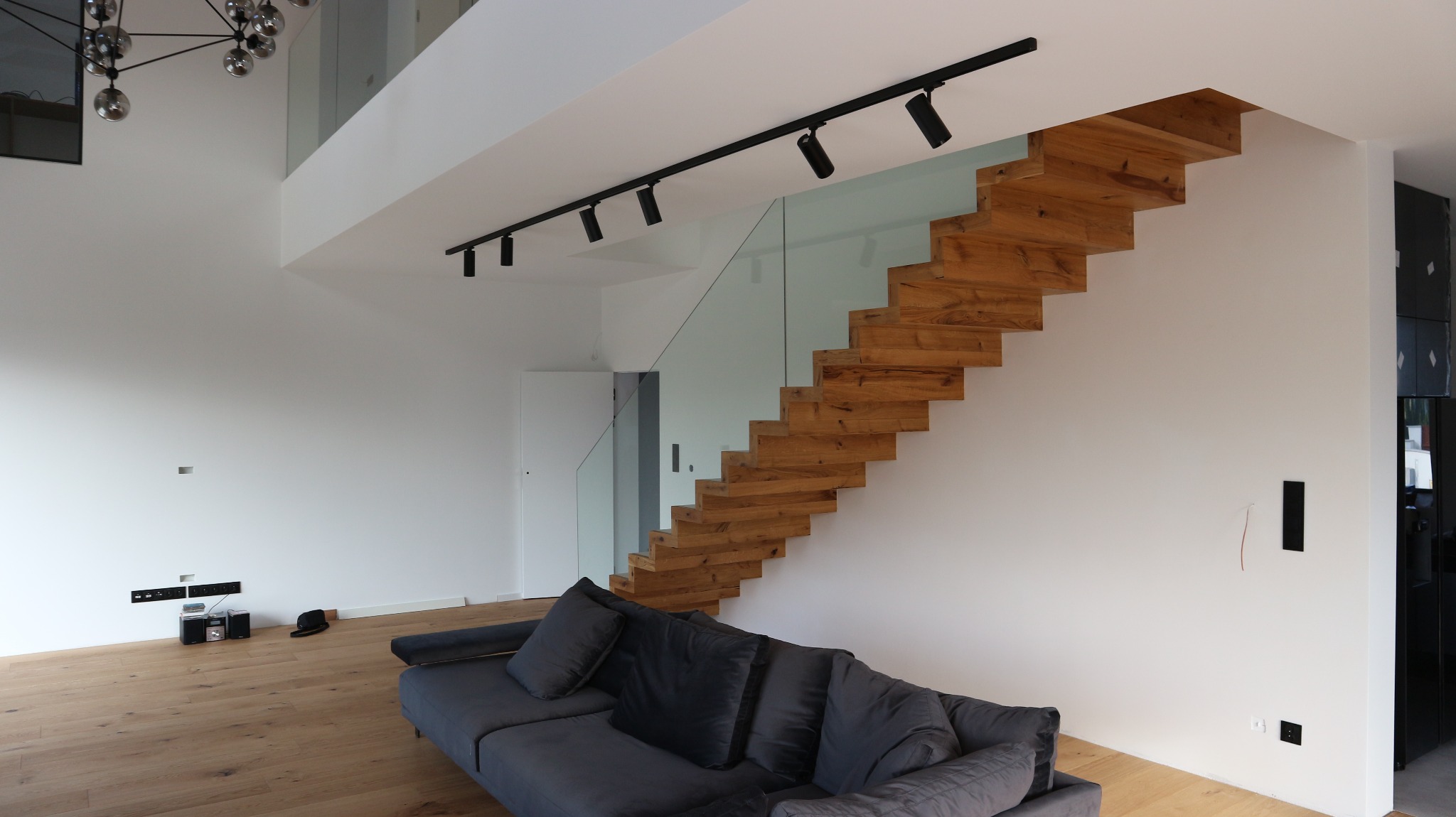 Nowoczesne wnętrze z pływającymi schodami z jasnego drewna, zabezpieczonymi szklaną balustradą, oświetlone reflektorami na szynie, z szarą sofą w minimalistycznym salonie.