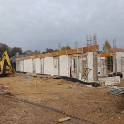 Budowa domu jednorodzinnego w trakcie wznoszenia ścian z betonu komórkowego. Widoczny żółty koparka i zbrojenia. Pochmurne niebo w tle.