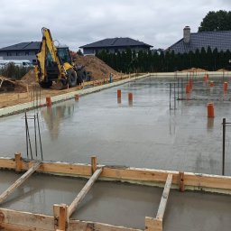 Zalany fundament pod budowę domu jednorodzinnego. Widoczne szalunki, wystające pręty zbrojeniowe i rury. W tle koparka i hałda ziemi.