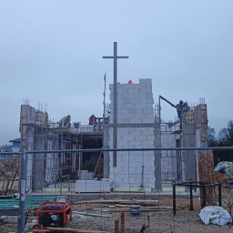 Firma Usługowa Łukasz Przyłęcki - Budowa kościoła w toku: murarze pracują na rusztowaniach, wznosząc ściany z białych bloczków. W tle widoczny krzyż i pochmurne niebo.