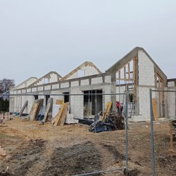 Firma Usługowa Łukasz Przyłęcki - Budowa domu z betonu komórkowego w stanie surowym otwartym, widoczne ściany, nadproża, drewniane szalunki okienne, teren budowy zabezpieczony siatką.