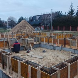 Firma Usługowa Łukasz Przyłęcki - Zbrojenie i szalunek fundamentów domu jednorodzinnego. Widoczny pracownik w trakcie pracy, narzędzia oraz hałda piasku w tle. Ujęcie z góry, pochmurny dzień.