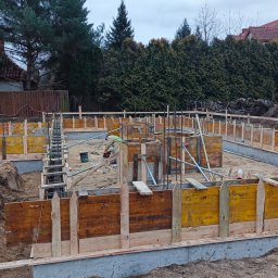 Firma Usługowa Łukasz Przyłęcki - Wykopane fundamenty z drewnianym szalunkiem i zbrojeniem, przygotowane do zalania betonem. W tle zieleń drzew i zabudowa jednorodzinna. Prace ziemne w toku.