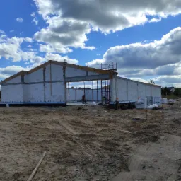 Wznoszenie ścian parterowego budynku z bloczków, widoczne drewniane elementy konstrukcji dachu, rusztowanie, materiały budowlane i dwóch pracowników w oddali na placu budowy, w tle błękitne niebo...