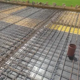 Zbrojenie fundamentu ułożone na folii z widocznymi dystansami i deskowaniem obwodowym, w tle trawnik i drzewa.