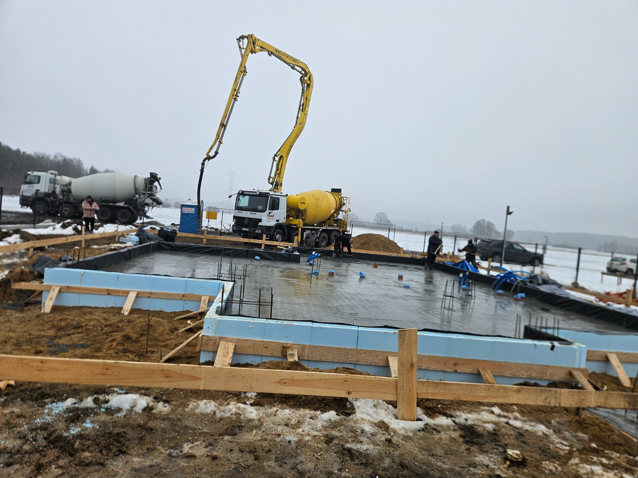 Wylewanie fundamentów domu jednorodzinnego w zimowej scenerii. Widoczny betonowóz z pompą, szalunki z izolacją i pracownicy budowlani.