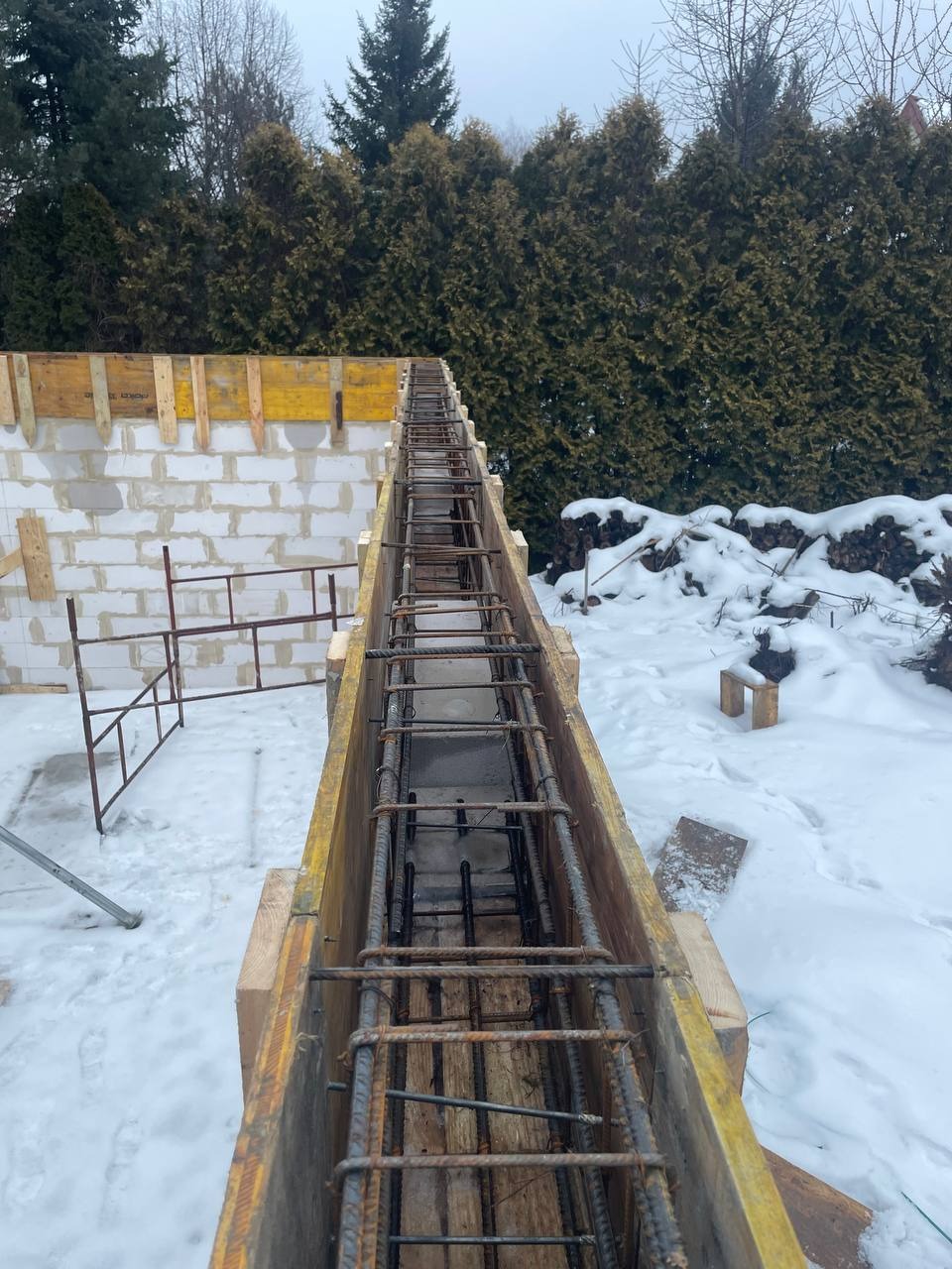 Zimowy widok na szalunek zbrojeniowy fundamentu, z widocznymi prętami i częściowo wymurowaną ścianą z bloczków, na tle ośnieżonego terenu i zielonych drzew.