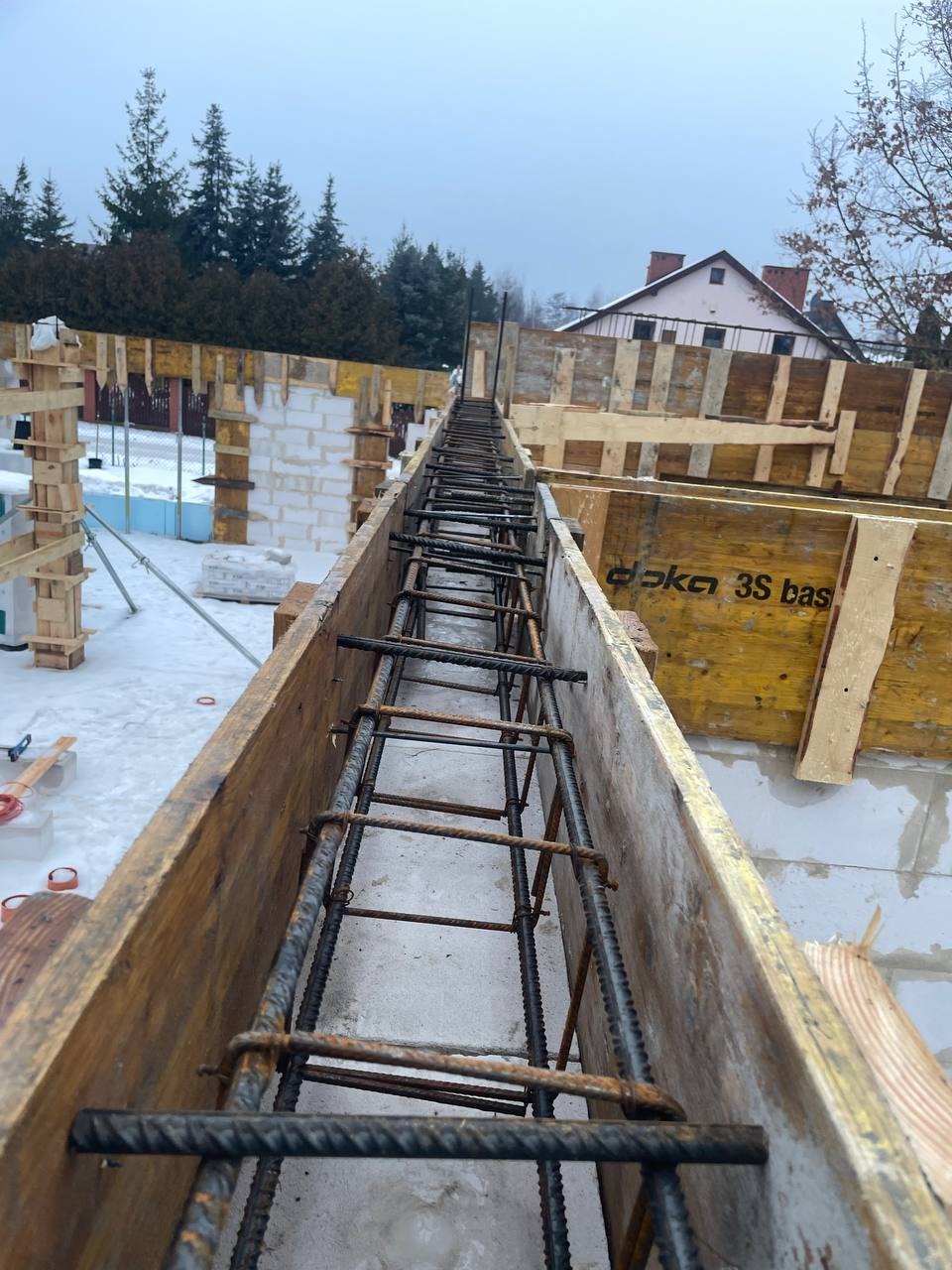 Zbrojenie w szalunku na fundamencie domu w zimowej scenerii. Widok z góry na stalowe pręty i drewniane deski, w tle budynek w trakcie budowy i ośnieżone drzewa.