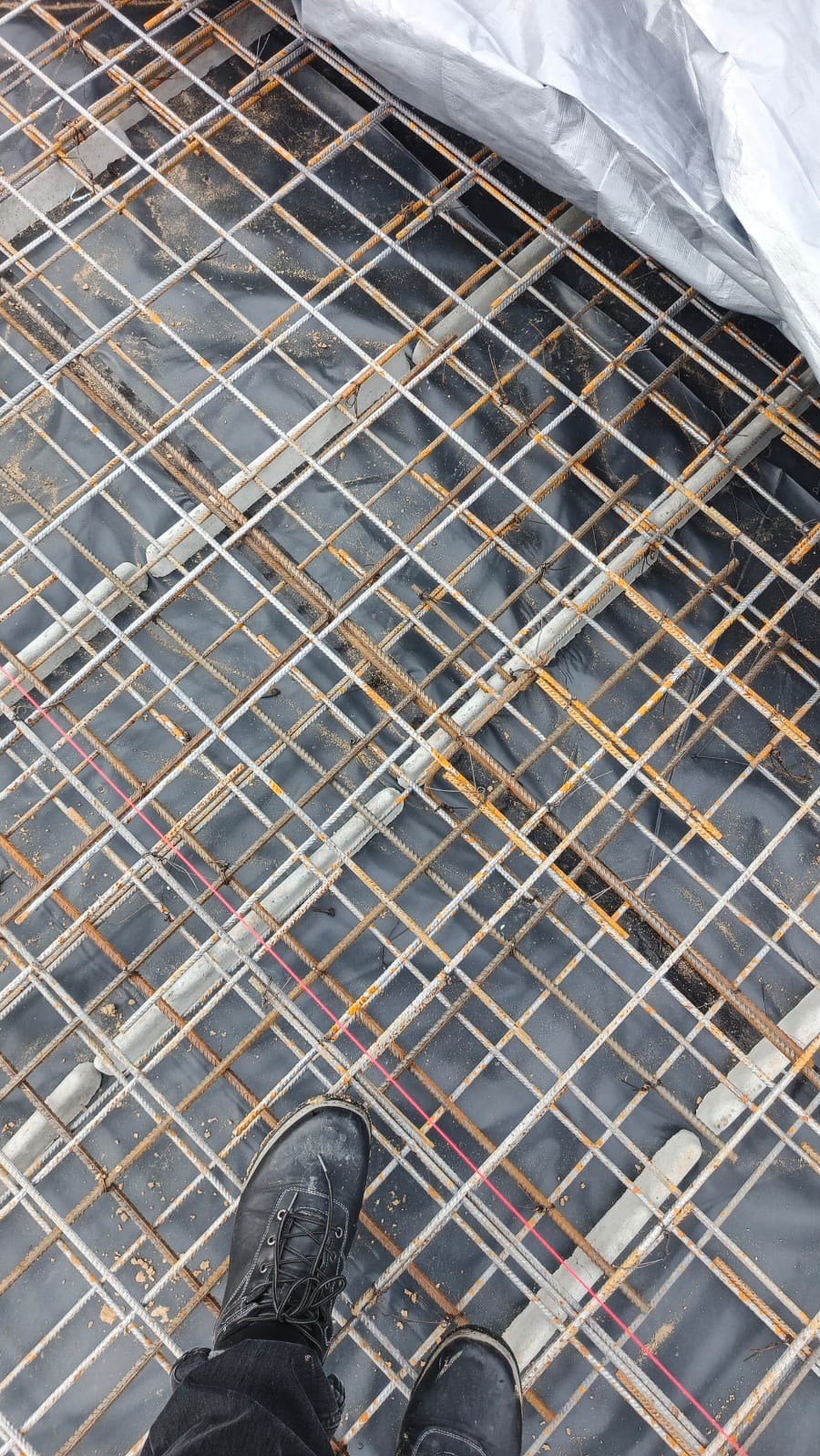 Zbrojenie fundamentów na czarnej folii, widoczne buty pracownika w lewym dolnym rogu. Siatka zbrojeniowa z prętów stalowych, częściowo przykryta folią ochronną.