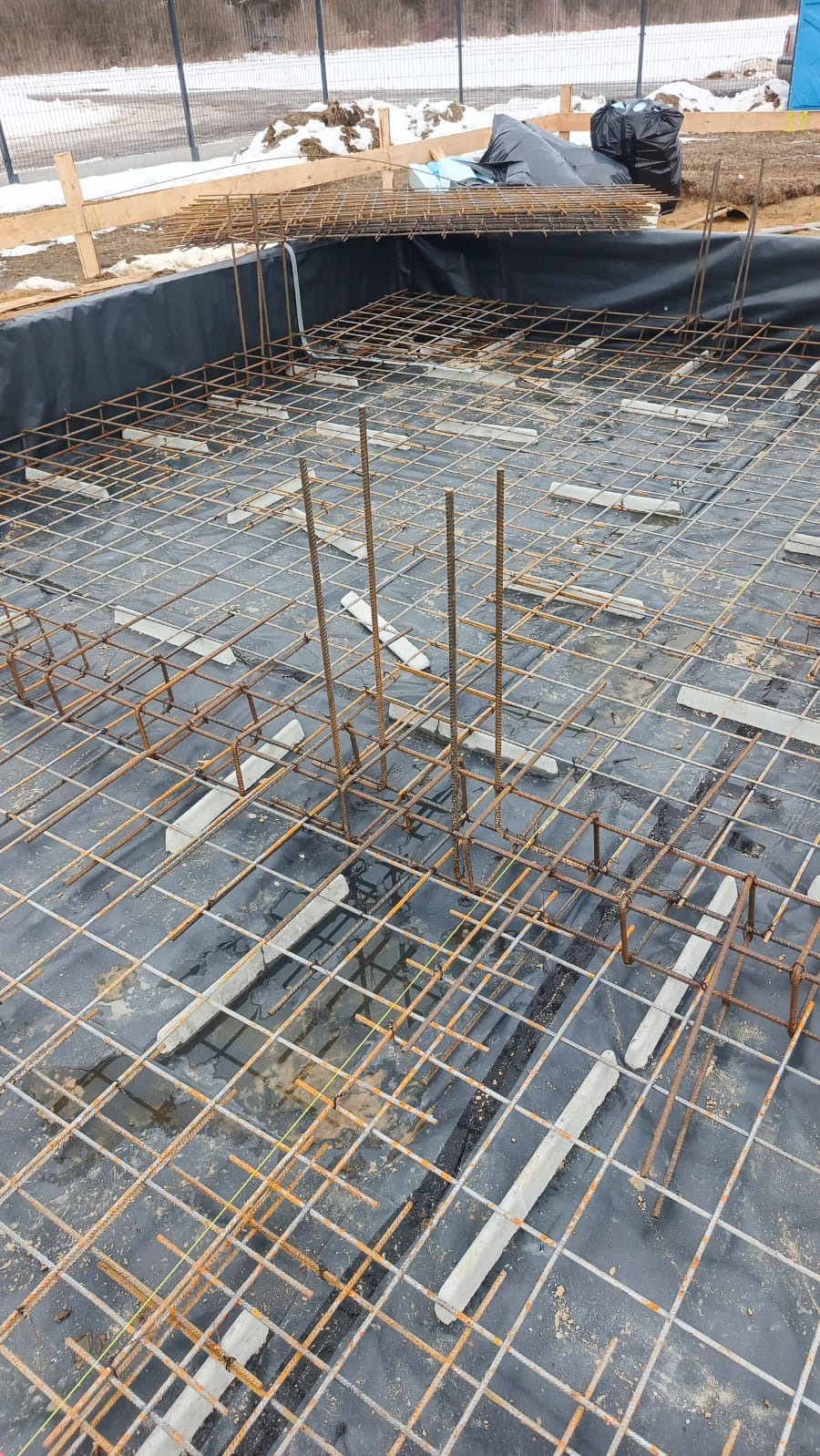 Zbrojenie fundamentu pod budynek, widoczne stalowe pręty tworzące siatkę, podkładki dystansowe i czarna folia izolacyjna. W tle teren budowy z resztkami śniegu.