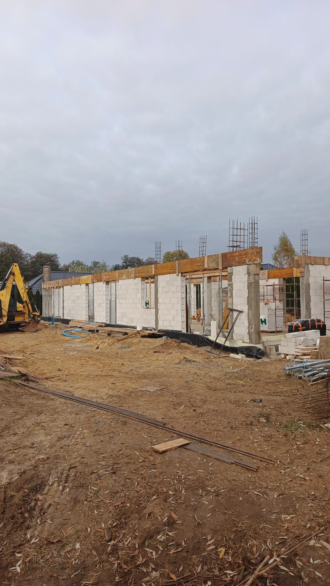 Budowa domu jednorodzinnego w trakcie wznoszenia ścian z betonu komórkowego. Widoczny żółty koparka i zbrojenia. Pochmurne niebo w tle.