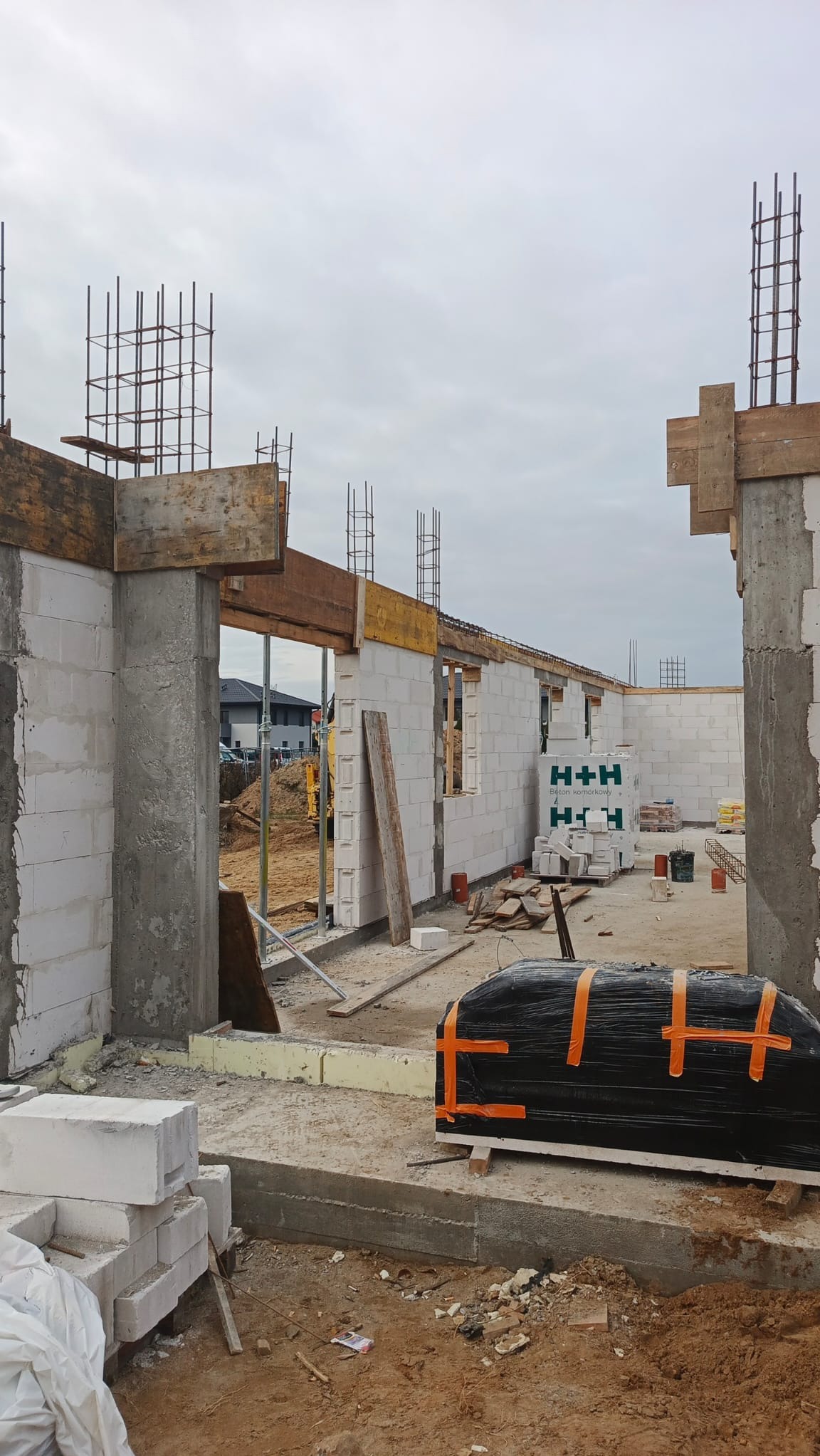Budowa domu w trakcie: ściany z betonu komórkowego H+H, zbrojenia pionowe, drewniane szalunki. Widoczny teren budowy i materiały budowlane.